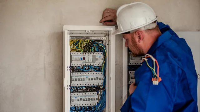 Stimmungsbild zum Beitrag: Es wird ein Elektriker im Blaumann und mit Helm vor einem geöffneten Stromkasten abgebildet.