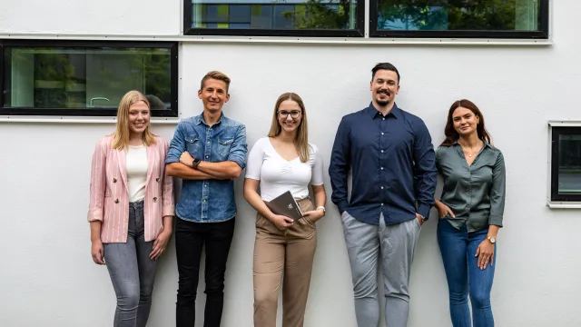 Stimmungsbild zum Beitrag: Es werden fünf stehende Auszubildende vor einer Hauswand abgebildet.