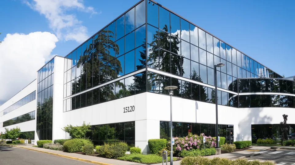 Bürogebäude mit großer Glassfassade, in der sich der Himmel spiegelt. Die Beete rund um das Gebäude sind mit Buchsbaum und weiteren Pflanzen angelegt.